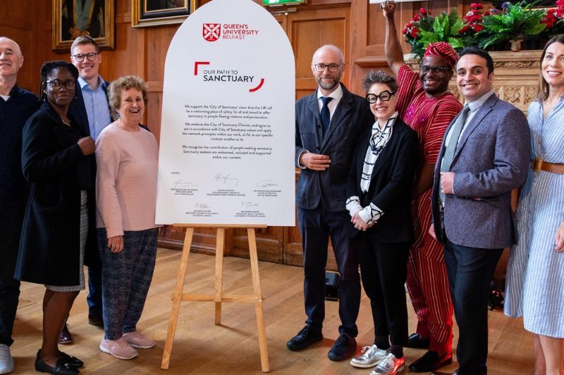 The Sanctuary Team: from L to R, John O’Neill, Tiwalade Olatunbosun, Raymond Miller, Sandra Griffiths, Maurice Macartney, Federica Ferrieri, Israel Eguaogie, Mehrshad Esfandiari, Aileen Cummins standing either side a large white cathedral window shaped board, with certification of University of Sanctuary status. Staff are wearing an array of suits, dresses, national dress and smart office wear.